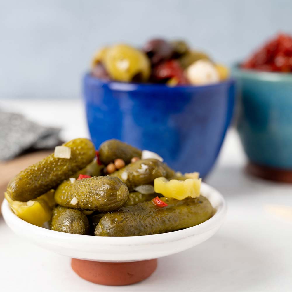 A bowl of cornichons in front of a bowl of antipasti olives and a bowl of sun-dried tomatoes that has been served out of 1kg foodservice pouches or tubs