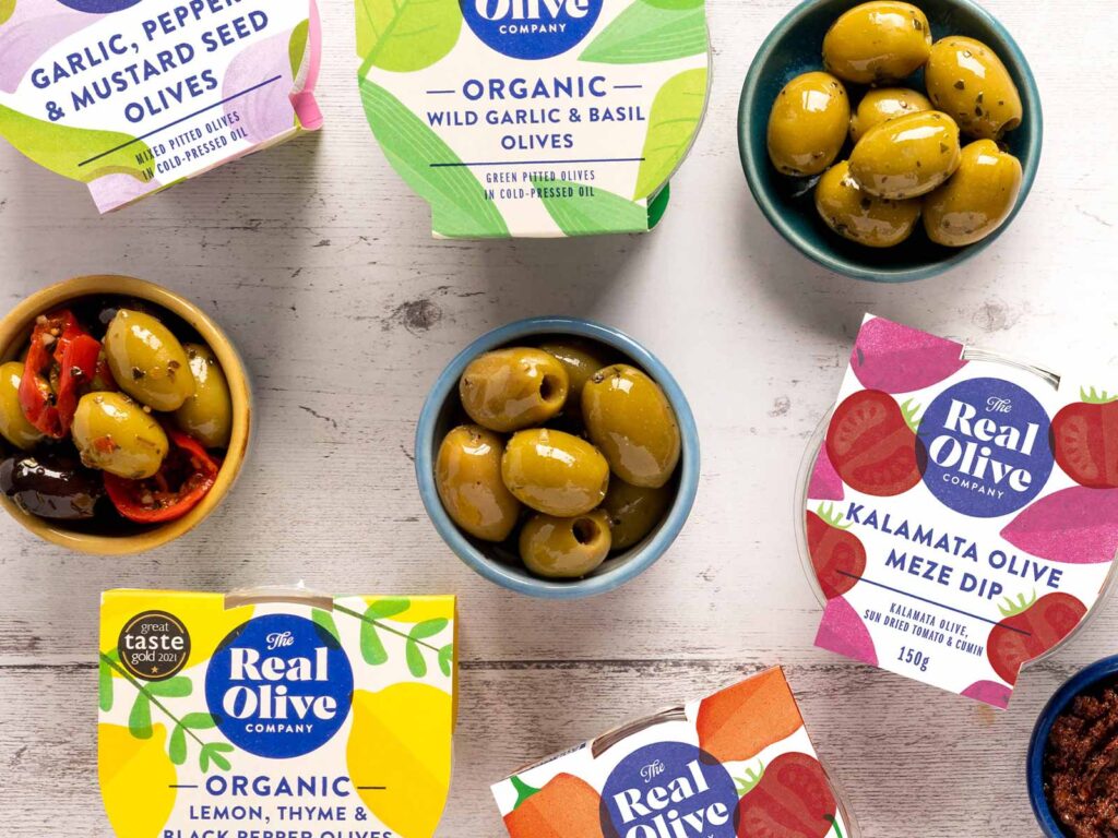 A selection of The Real Olive Company retail pots of olives and Kalamata olive meze dip in stand out packaging and bowls filled with the products.